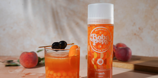 Boba Pop's peach-flavored drink with a glass of an old-fashioned cocktail and peaches on a wooden surface.