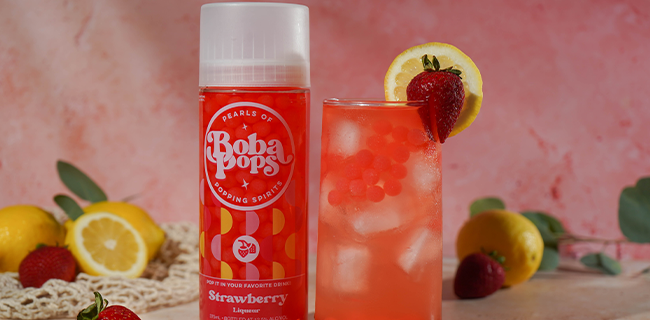 Boba Pops strawberry flavor bottle next to a glass of strawberry lemonade with fruit on a pink background