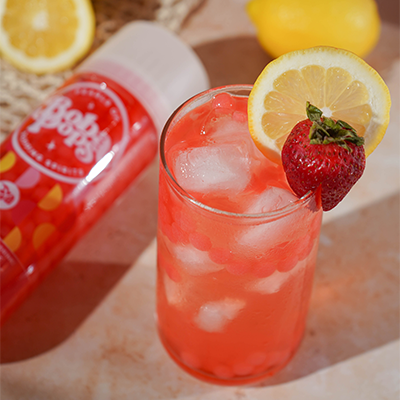 Glass of pink lemonade with a strawberry garnish, next to a bottle of Boba POPS Strawberry flavor and lemons.